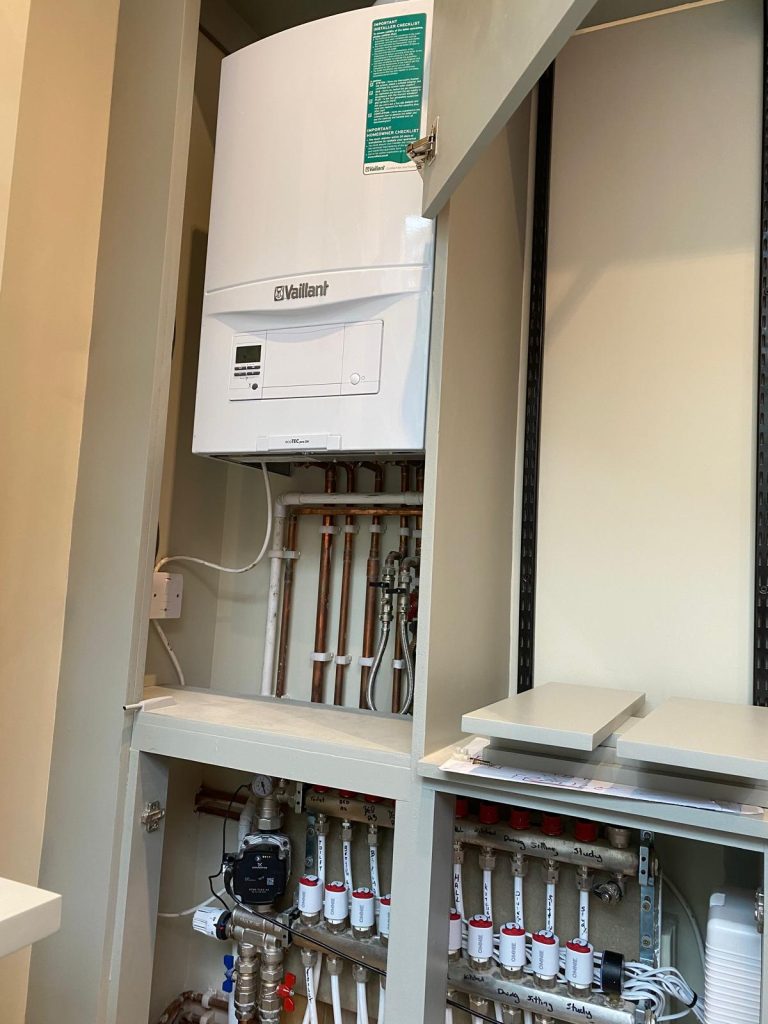 Vaillant boiler installed above a neat underfloor heating manifold system inside a cabinet, showing well-organised copper pipework and valves.