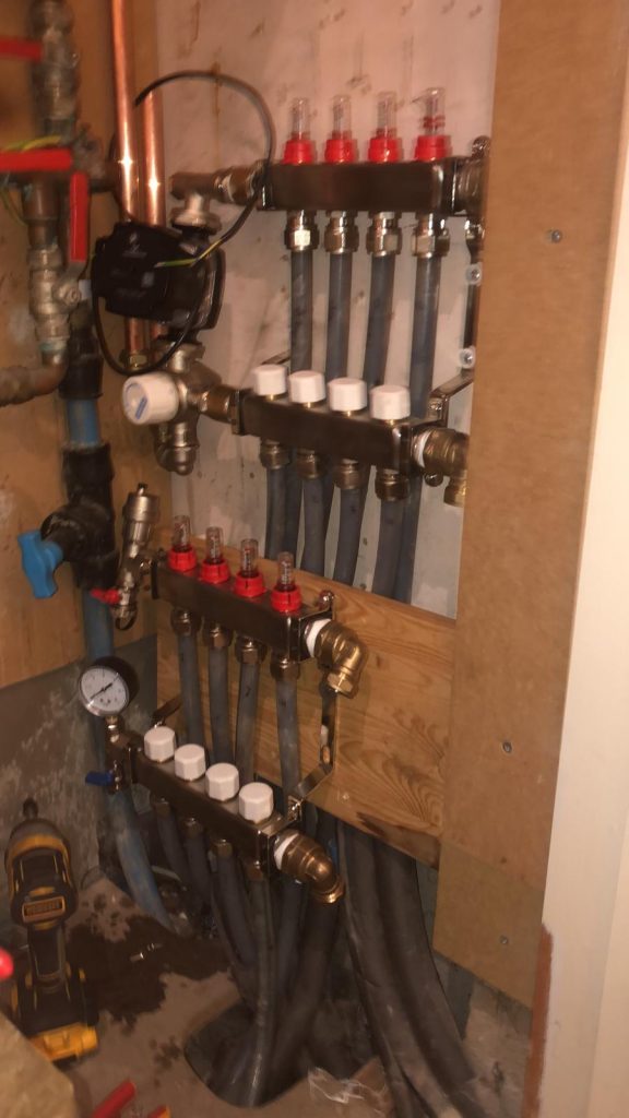 Vertical underfloor heating manifold installation featuring multiple zones with grey pipework, flow gauges, actuators, and a Grundfos circulation pump in a tight service area.