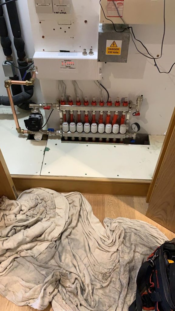 Underfloor heating manifold with multiple valves and connected pipes inside a cabinet, alongside an electrical control unit, with a protective dust sheet on the floor.